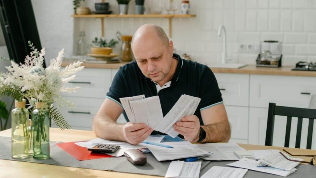 A balding man in a dark polo shirt is sitting at a kitchen table, intensely reviewing several papers.