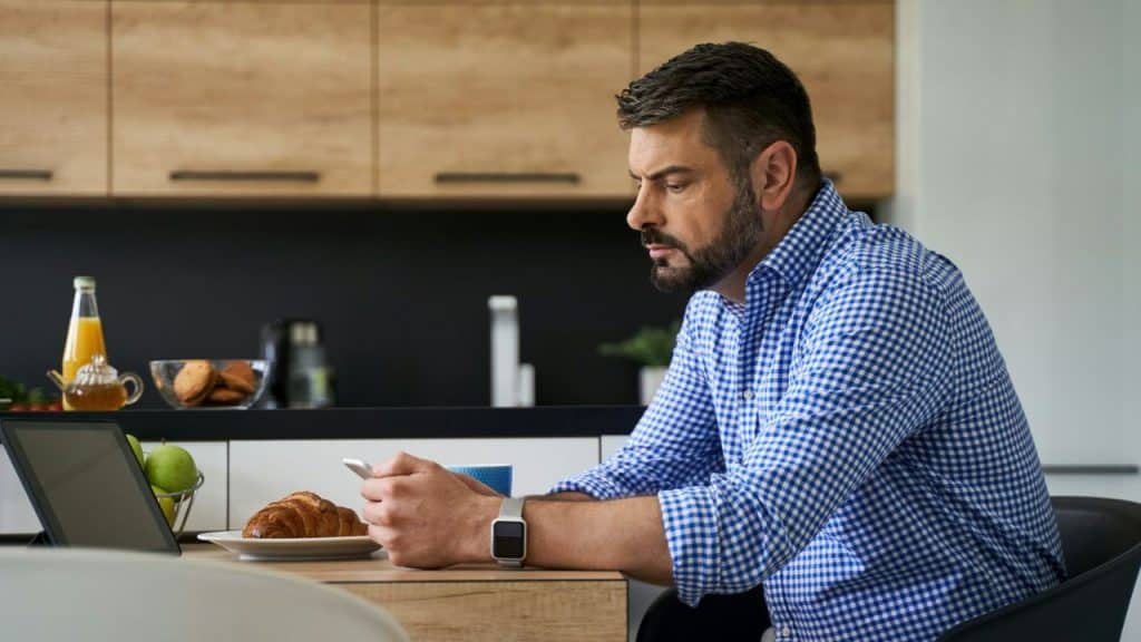 Man in a blue plaid shirt at a kitchen table using his smartphone.