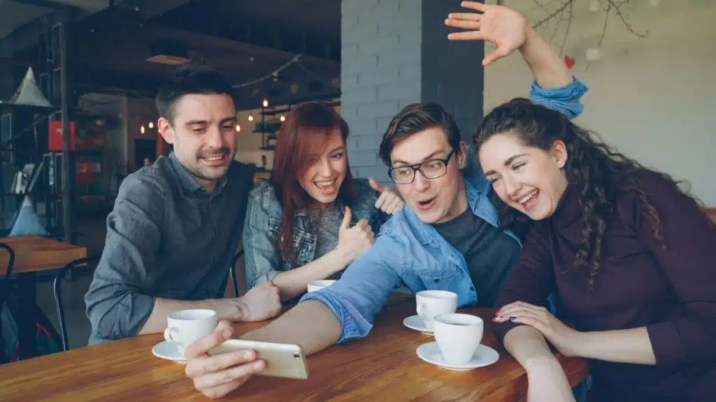 Four happy young adults are sitting at a table in a cafe, taking a selfie with a smartphone.