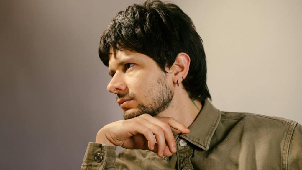 Close-up profile of a man with dark hair and a beard, resting his chin on his hand.