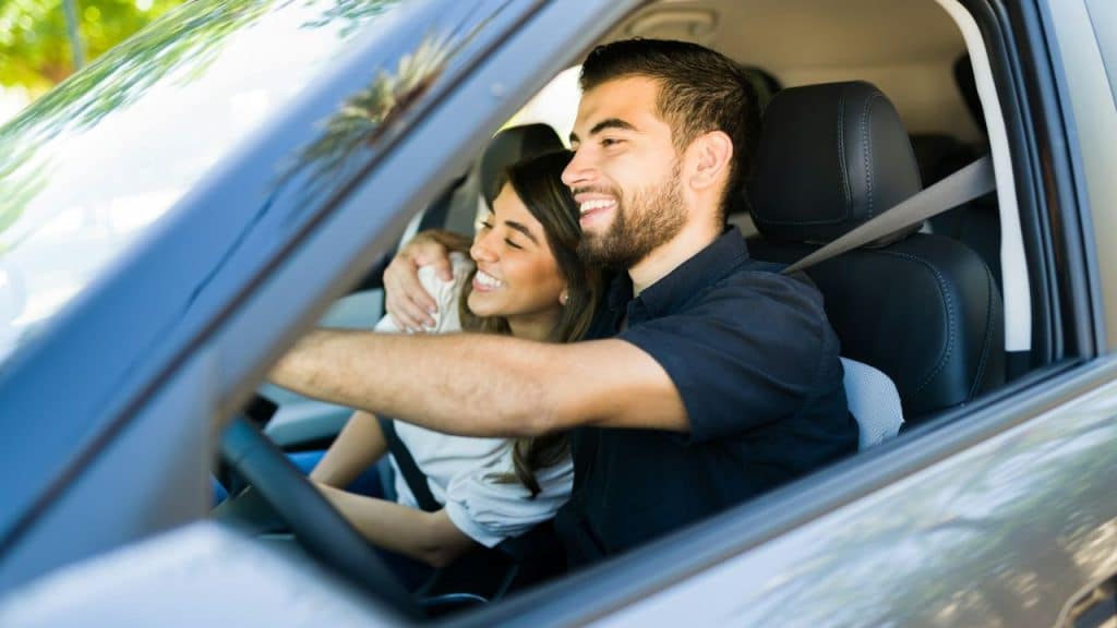 Smiling man drives a car with his arm around a woman in the passenger seat.