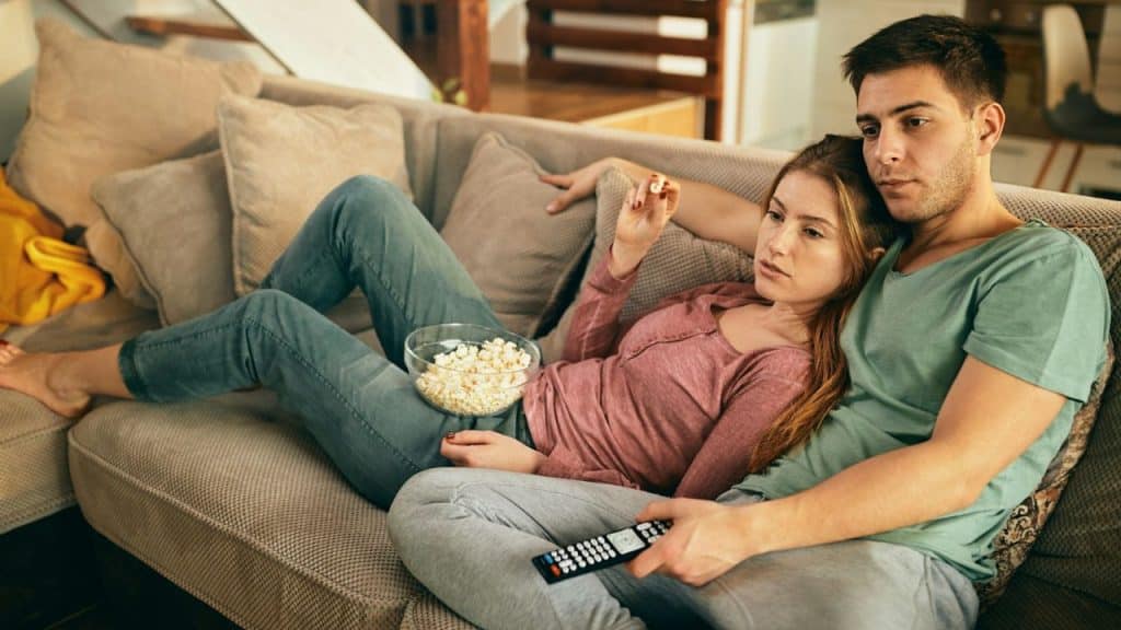 A man and woman are lying on a couch, watching TV with a remote and a bowl of popcorn.