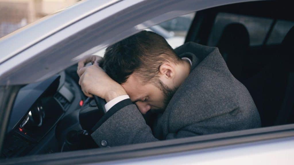Man in a gray coat resting his head on his arms on a car's steering wheel.