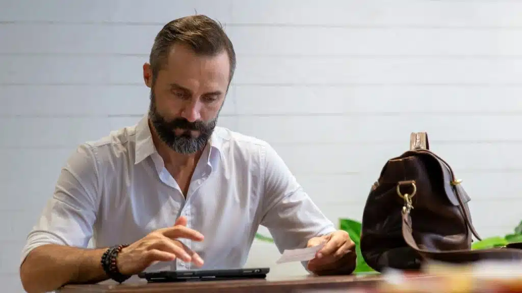 Bearded man in white shirt looking at a tablet and receipt with a brown bag nearby.