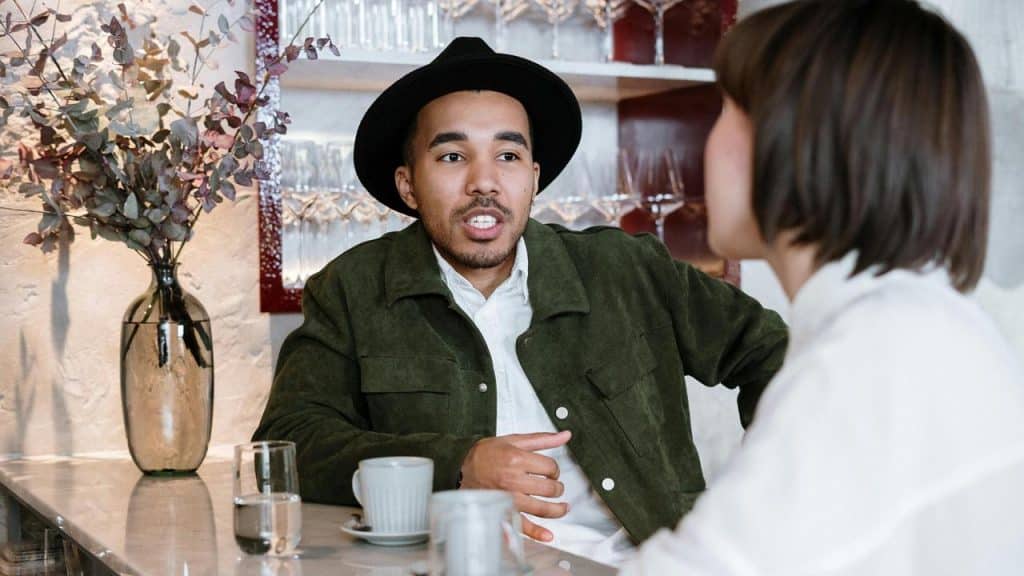 A man wearing a hat talking with a woman at a café table.