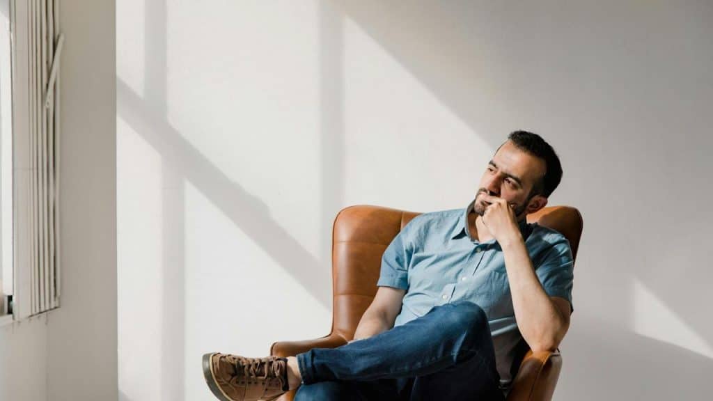 A man sitting on a chair in sunlight, resting his chin on his hand while thinking.