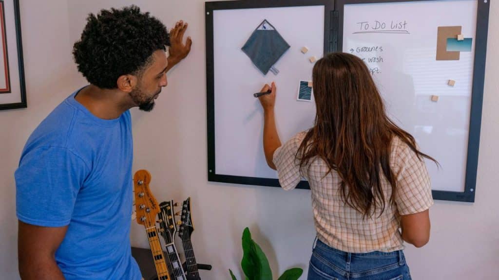 A woman with long brown hair writes on a white magnetic whiteboard while a bearded man in a blue shirt looks over her shoulder.