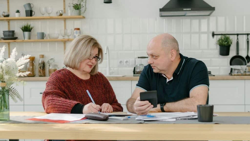 A man holding a calculator looks at a woman writing at a table in a bright kitchen.