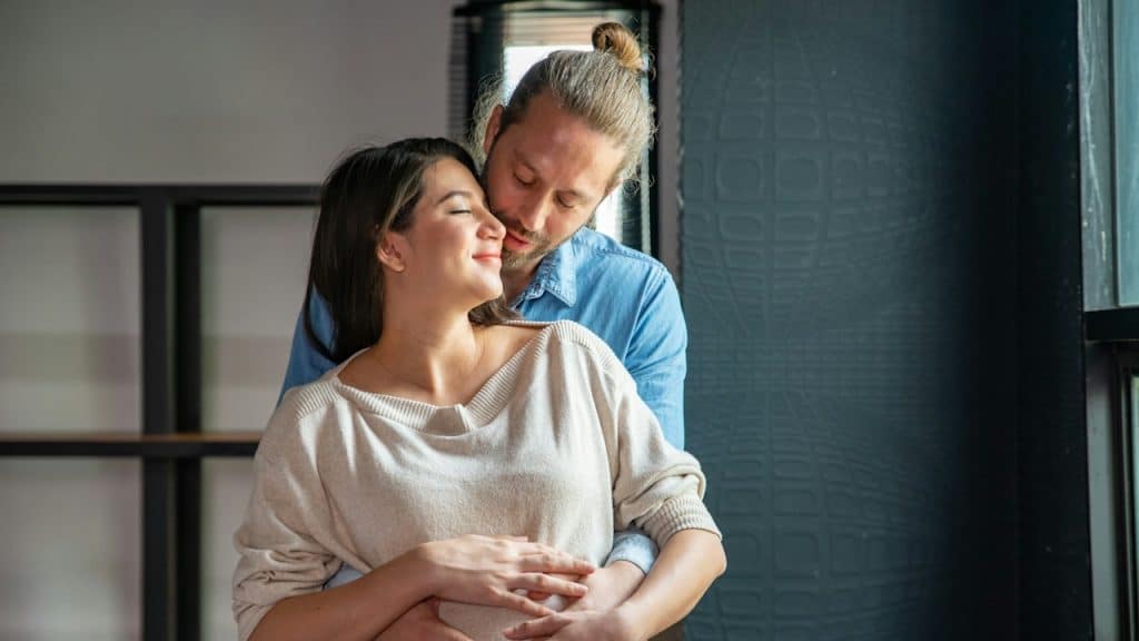 A smiling couple embraces indoors, with the man hugging the woman from behind and resting his chin near her shoulder.