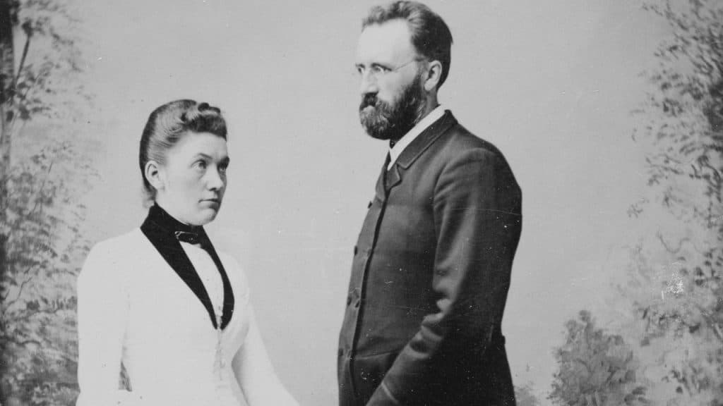 A black and white portrait of a man with a beard and glasses next to a woman wearing a white dress and high black collar.