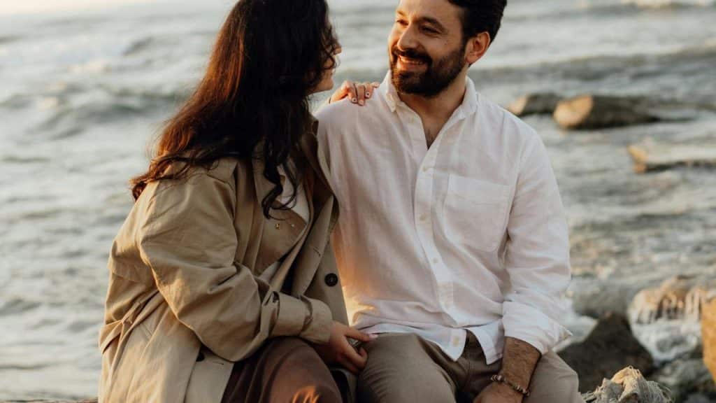 A couple sitting by the sea and smiling at each other.