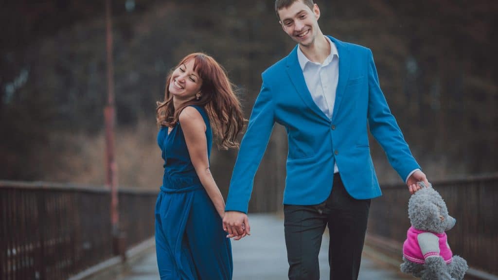 A man and a woman holding hands and walking with a teddy bear.