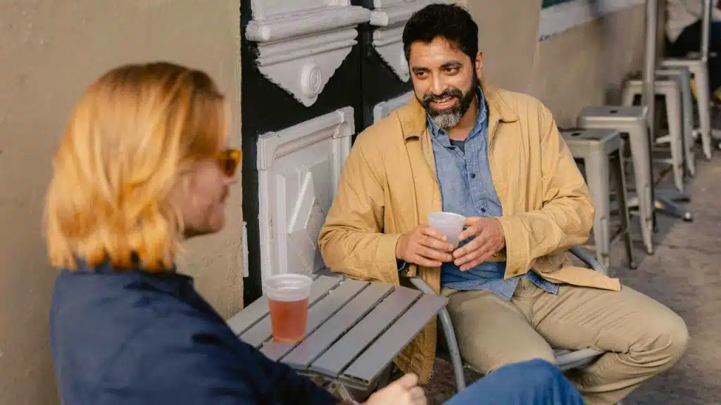 Bearded man and a blonde person sitting at an outdoor table, talking and holding cups.