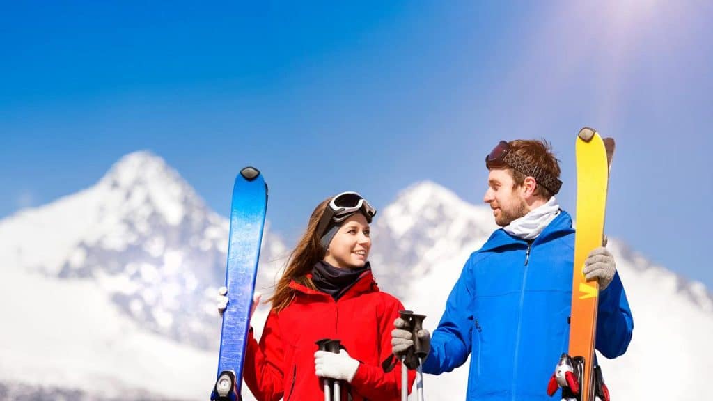 A couple holding skis and smiling with snowy mountains in the background.