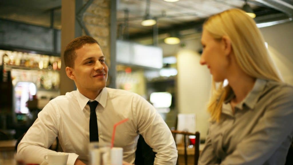 A young man in a white shirt and black tie smiles and looks across a table at a blonde woman in a gray shirt in a softly lit restaurant or cafe.