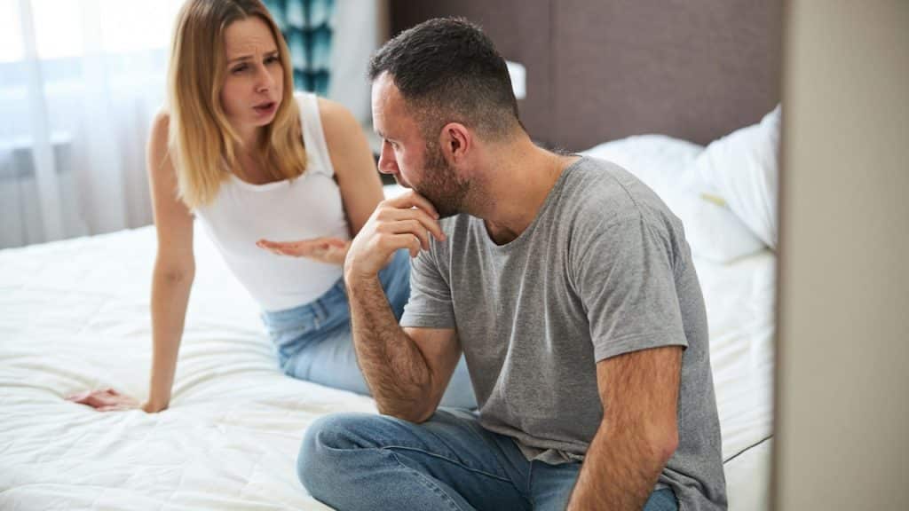 A displeased blonde lady sitting on a bed and talking with her husband.