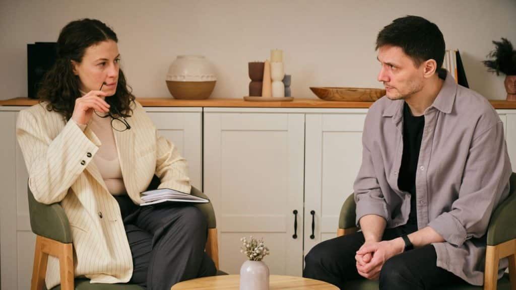 A woman with glasses and a man sit facing each other in armchairs in an indoor setting.