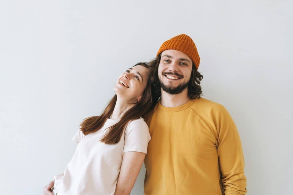 The couple standing in front of the gray wall is smiling.