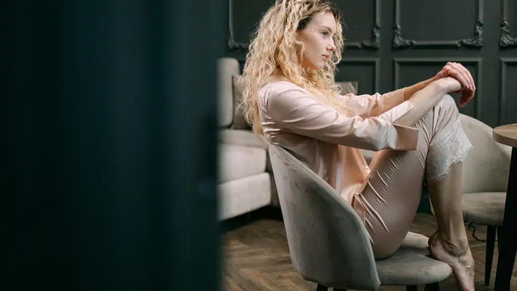 A woman in loungewear sits curled up on a chair, looking thoughtful.