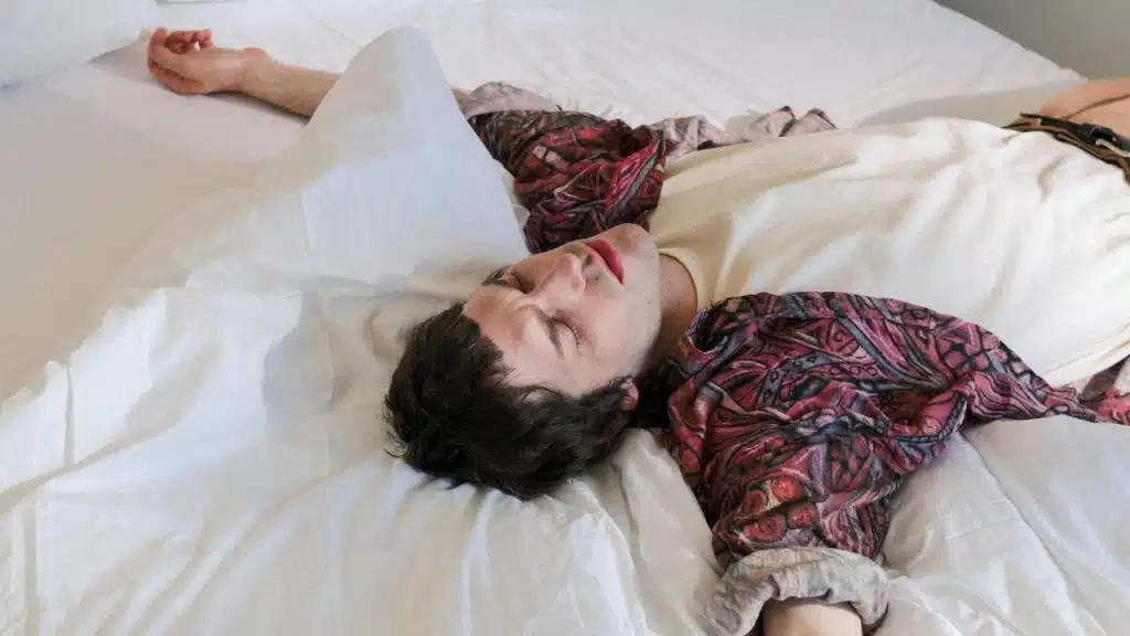A person lying on a bed with arms spread out, wearing a patterned shirt and white shirt.