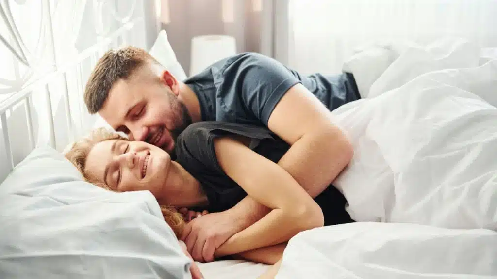 A smiling man is embracing a smiling woman who is lying on her back in a bed.