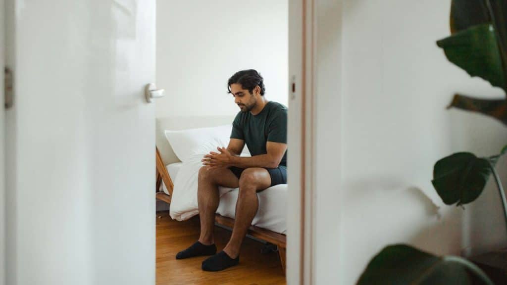 A man in a green shirt is sitting on the edge of a bed, looking down sadly.
