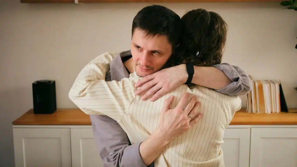 A man is embracing a woman from behind, looking over her shoulder with a concerned expression.