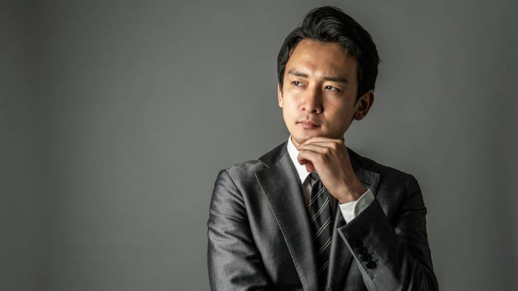 A suited, confident man thinking while holding his hand up to his face. 