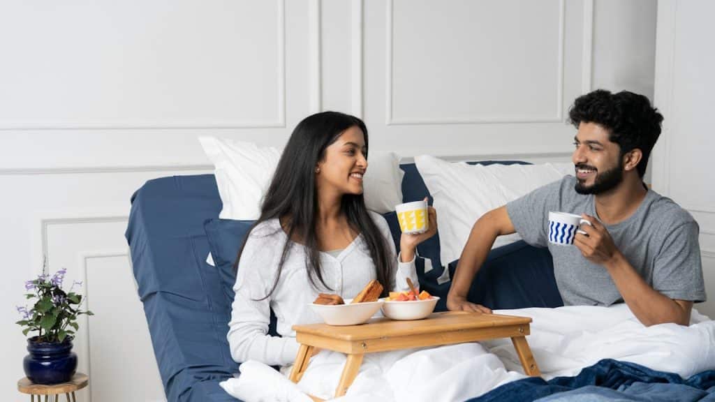 A smiling couple sits up in bed, enjoying breakfast in bed on a wooden tray with coffee cups.