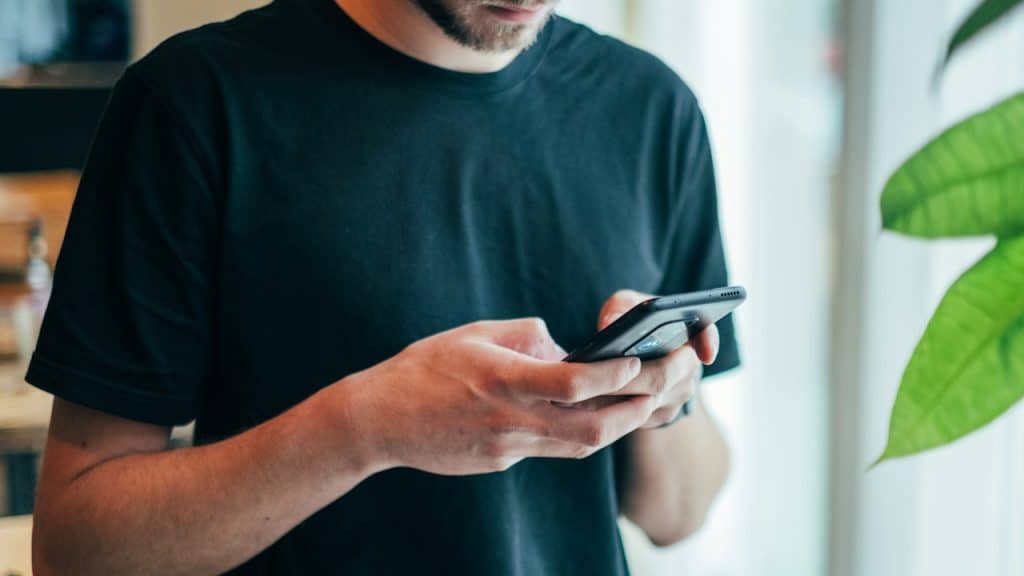 A man in a black T-shirt using a smartphone, with a green plant visible on the right.