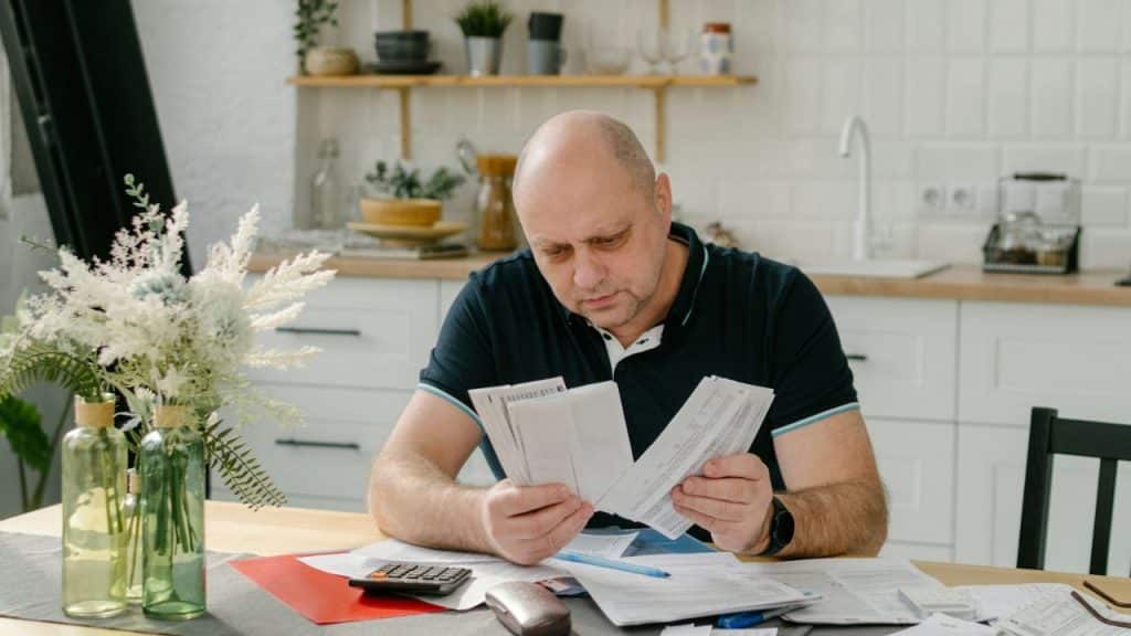 A balding man sits at a kitchen table, looking worriedly at several papers and bills.