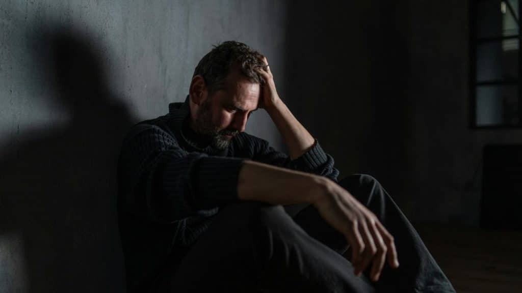 A bearded man sits in a dark, empty room with his head in his hand, looking down in distress.