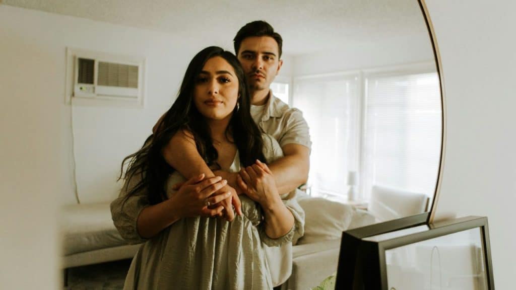 A serious couple embraces, reflected in a mirror inside a bright, modern room.