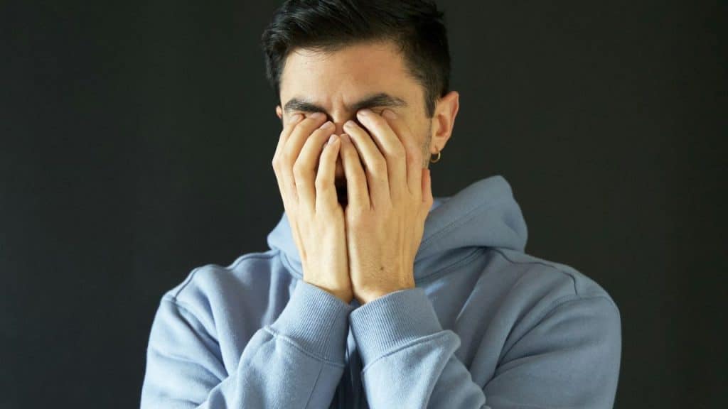 Man in a blue hoodie covering his face with his hands, against a dark background.