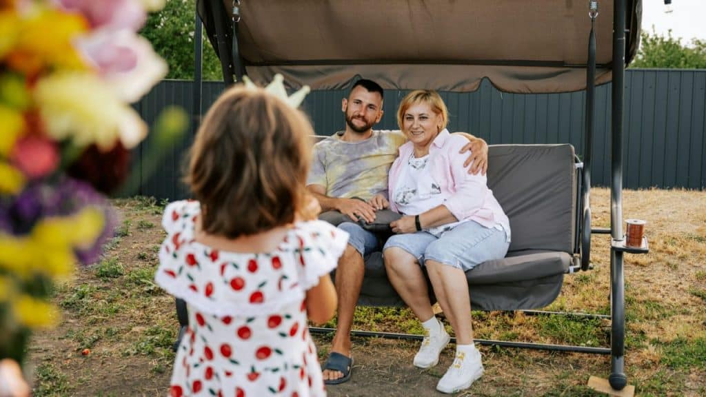 Man and woman seated on an outdoor swing looking at a young girl standing in the foreground.
