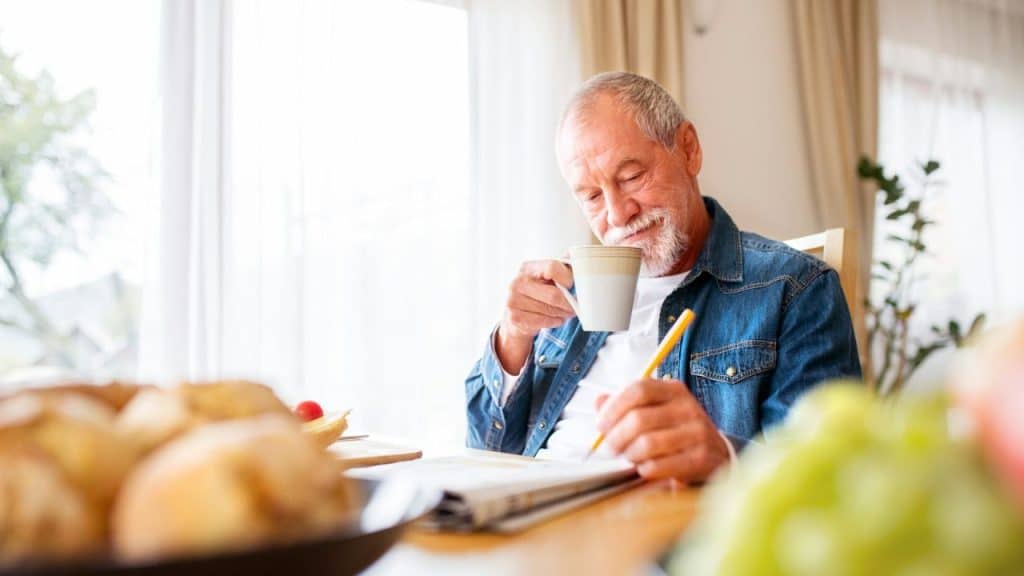 Senior man in a denim shirt sips coffee and writes at a table.