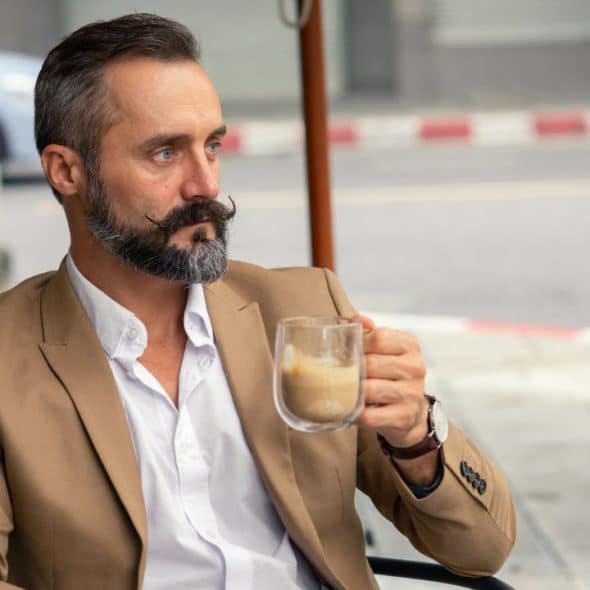 A stylish man with a mustache and beard is drinking coffee outdoors.