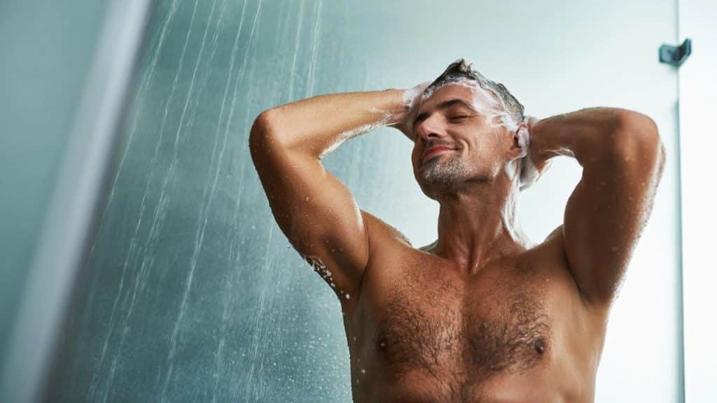 A smiling, muscular man is washing his hair in a shower with his eyes closed.