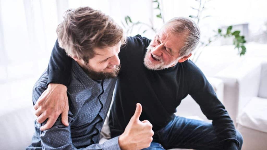 Older man hugs younger man giving a thumbs-up on a white couch.