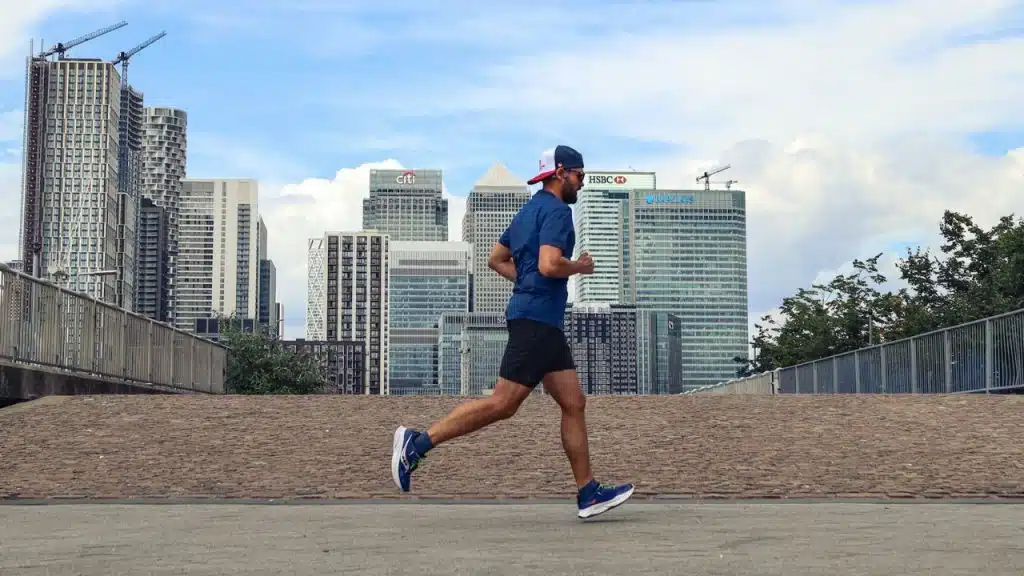 Man in shorts and a t-shirt runs across a paved area with a city skyline behind him.