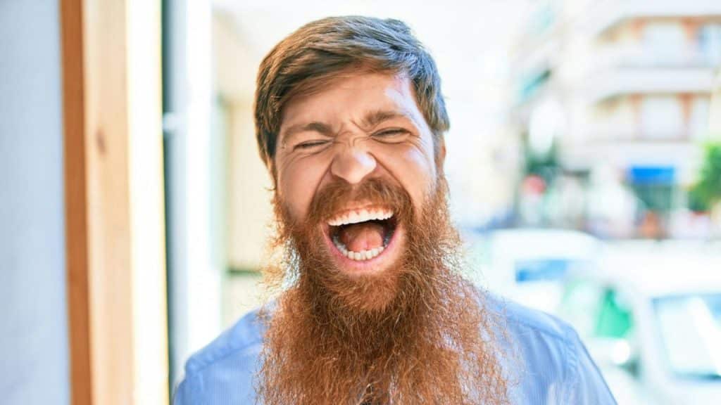 A man with a large red beard is yelling loudly with his eyes squeezed shut.