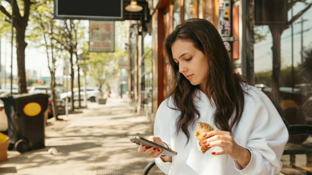 A woman sitting outside holding a phone in one hand and a piece of bread in the other.