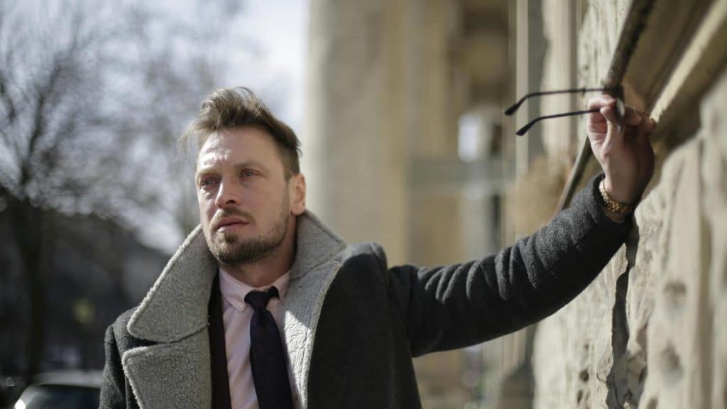 A man in a coat and tie holding his glasses against a wall.