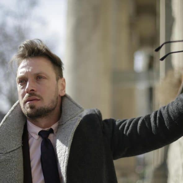 A man in a coat and tie holding his glasses against a wall.