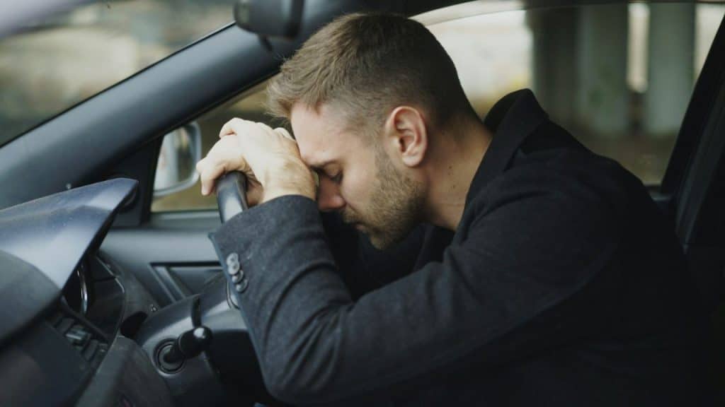 Close-up of a man in a dark coat slumped over his car's steering wheel.
