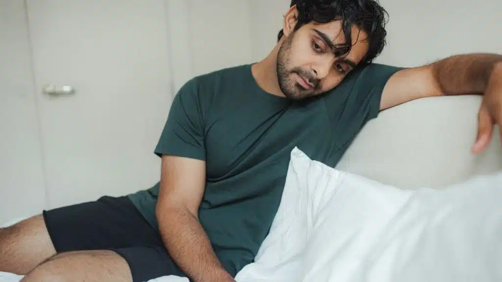 Young man in a dark green t-shirt sitting on a bed, looking down sadly.