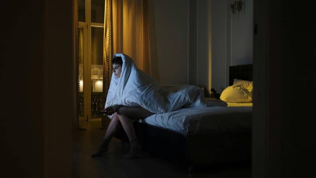 A person sitting on a bed wrapped in a blanket at night.