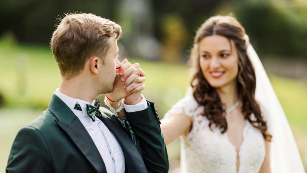 A groom kissing the bride’s hand while she smiles at him outdoors.