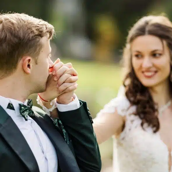 A groom kissing the bride’s hand while she smiles at him outdoors.