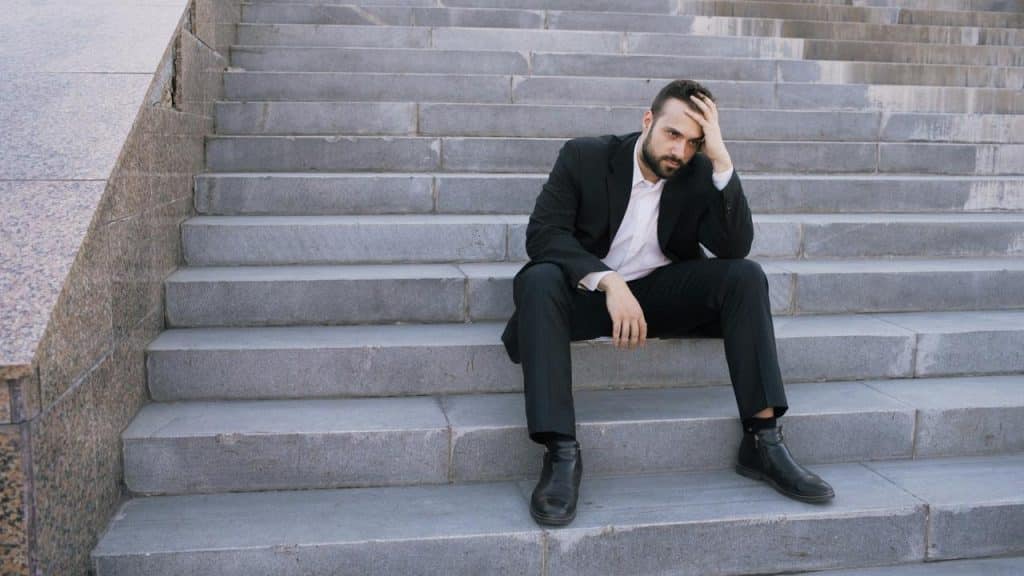 A man in a suit sitting on outdoor steps with his hand on his head looking stressed.